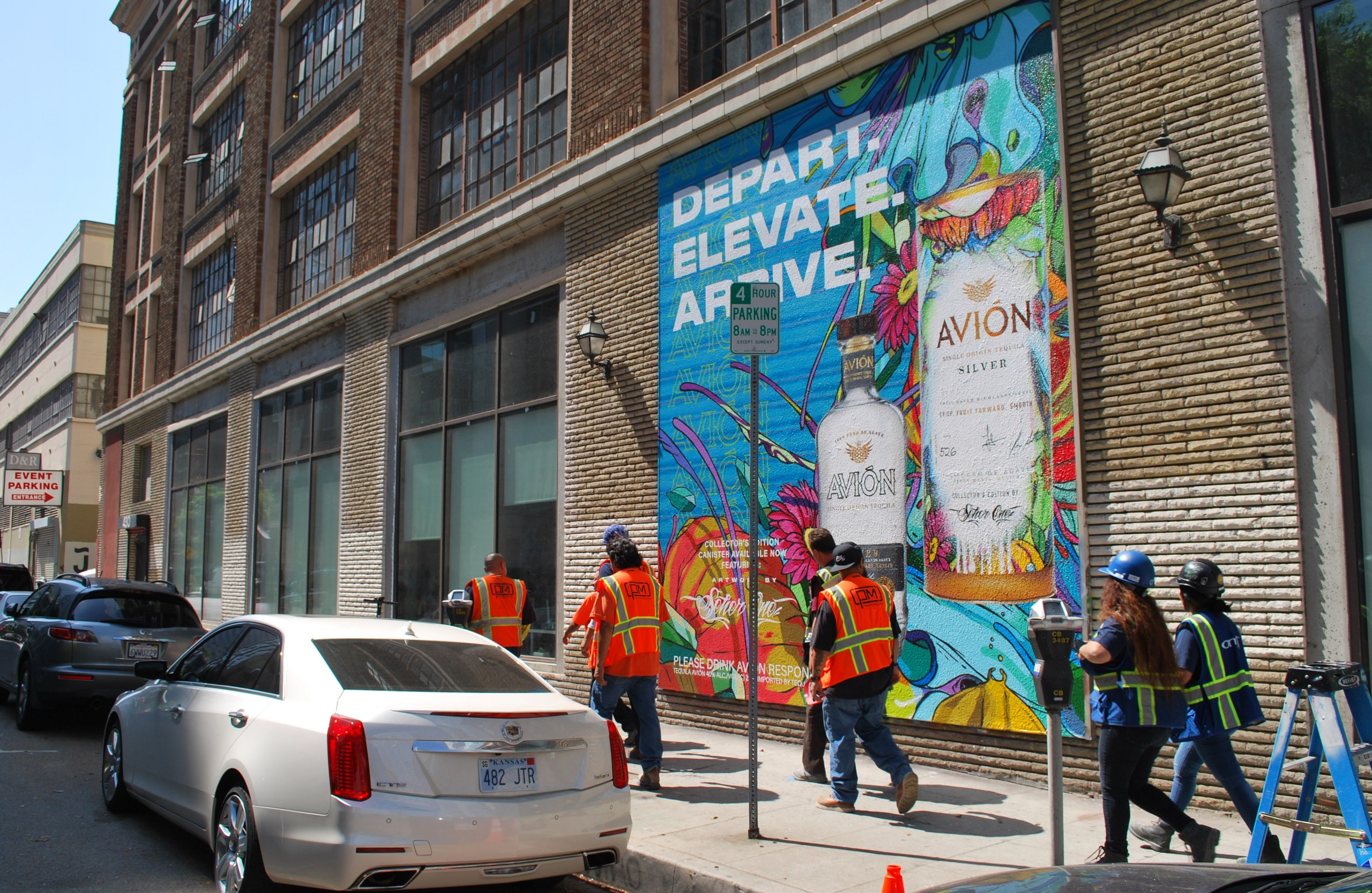Workers install a large Avión Tequila mural on a brick building along Hope Street in Los Angeles, featuring colorful artwork and oversized bottle graphics.