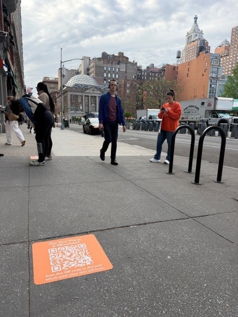 Orange sidewalk decal with a QR code installed on a New York City sidewalk near Union Square, with three pedestrians walking past and one looking at her phone.