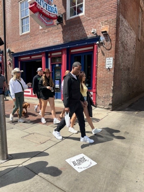 Group of young adults walking on a sunny sidewalk past a brick storefront with blue and red trim.