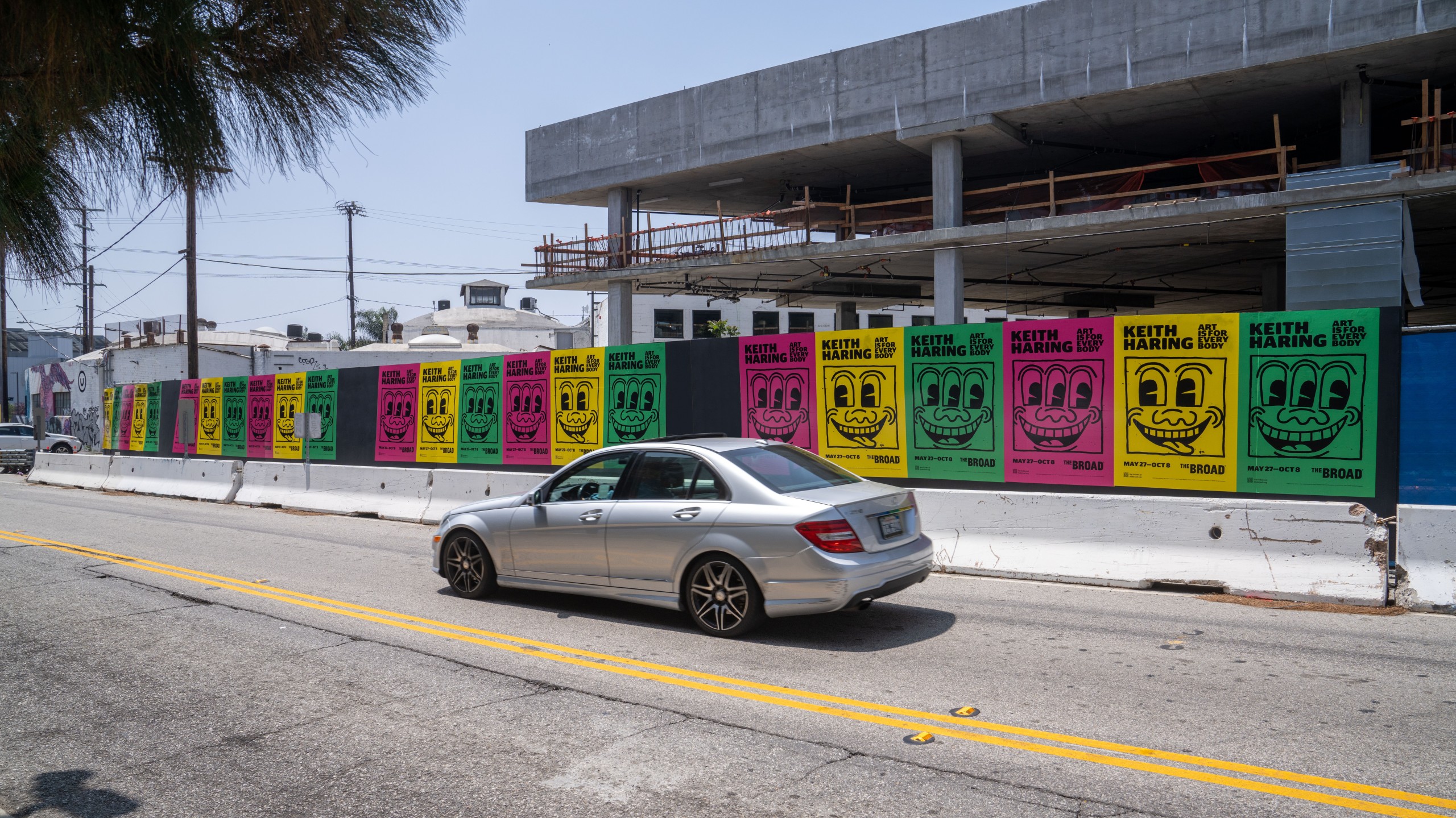 Colorful Keith Haring posters for The Broad museum’s “Art Is for Everybody” exhibit pasted along a construction wall in Los Angeles, with a silver car driving by.