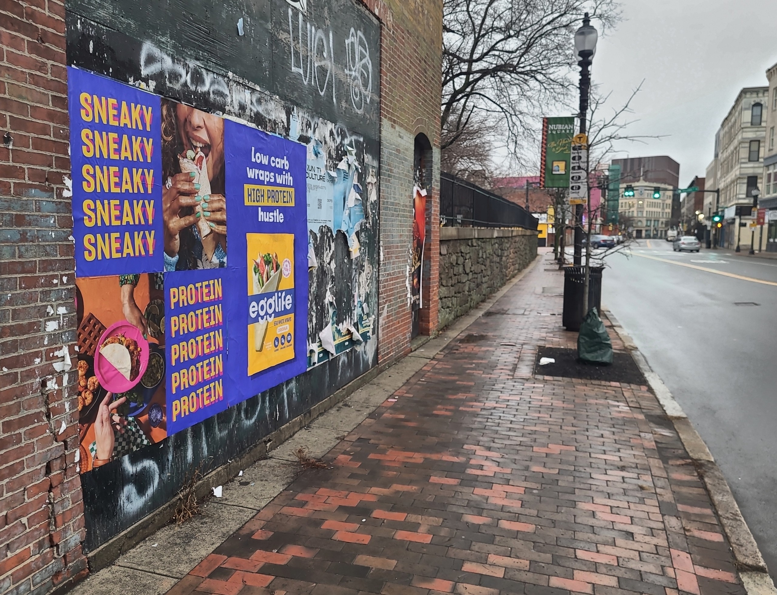 Wild Posting® posters for Egglife egg white wraps displayed on a brick wall in Boston, Massachusetts, along a busy sidewalk.