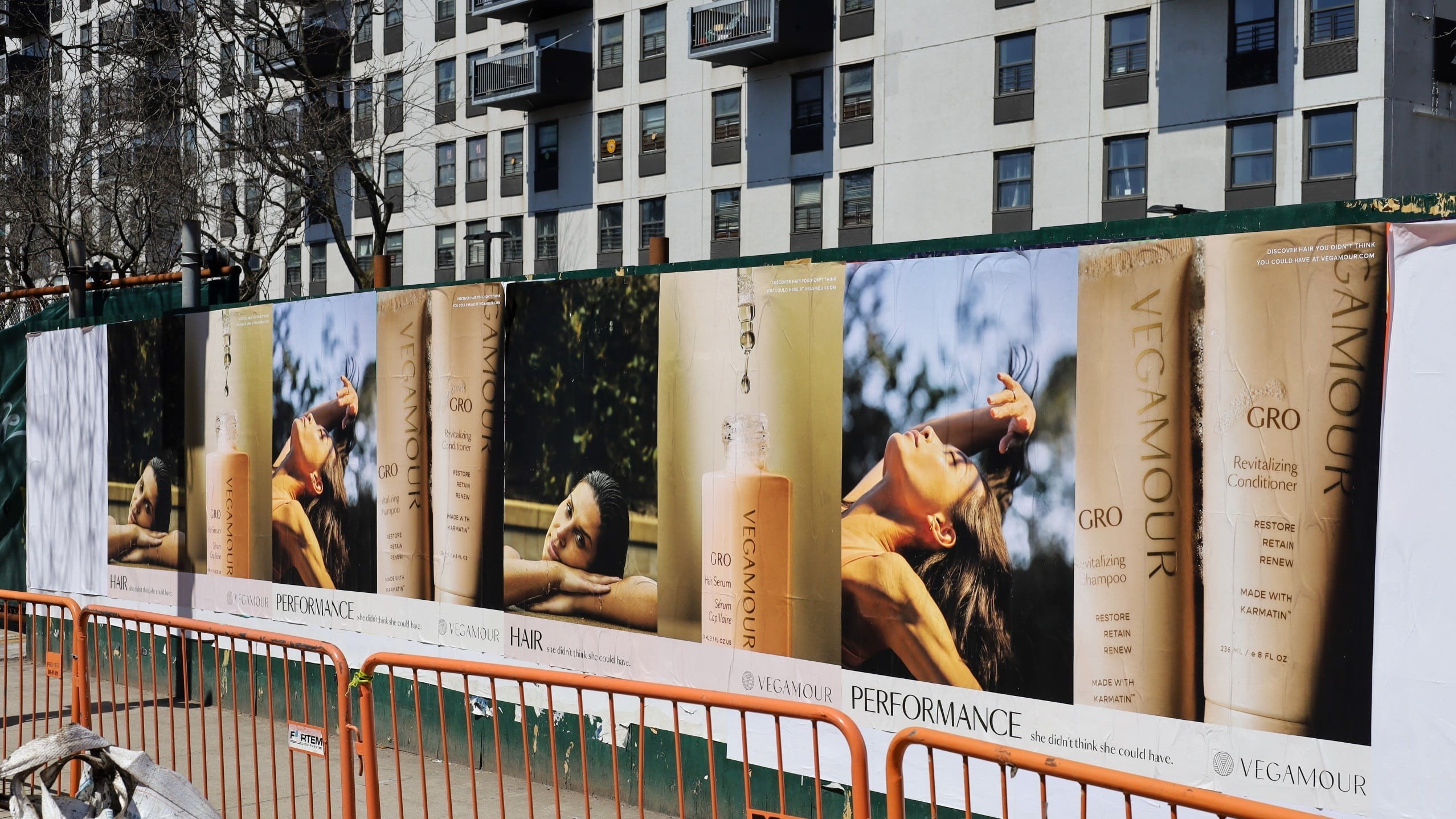 Large VEGAMOUR hair-care Wild Posting® installation on a construction wall in Williamsburg, Brooklyn, with beauty product images and models displayed across multiple posters.