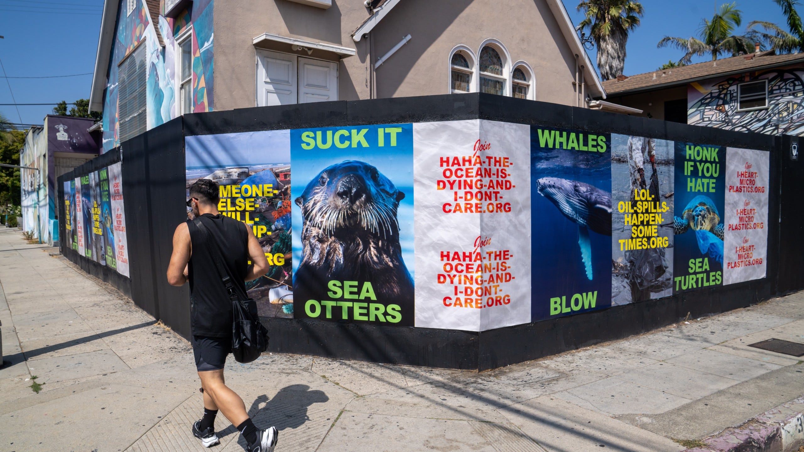 Wall covered in colorful Wild Posting® environmental posters with sea animals and anti-pollution slogans, located on a Venice Beach street corner as a pedestrian walks by.