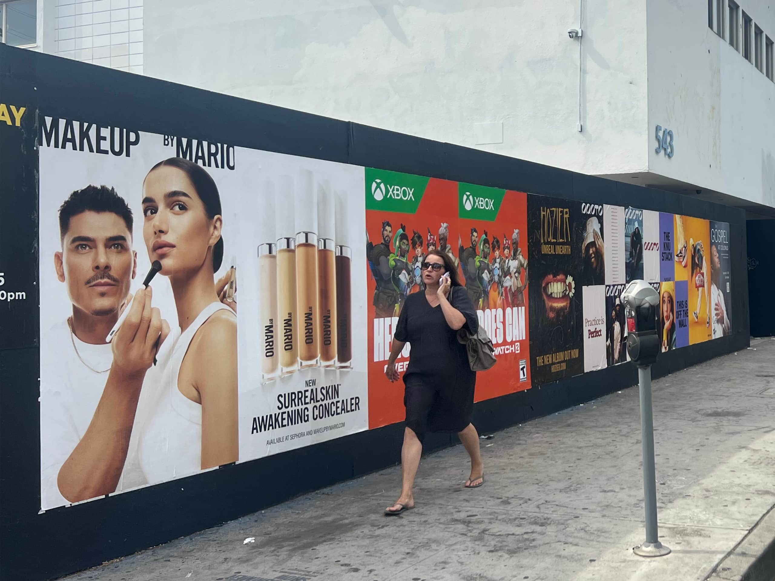 Wild Posting® posters for Makeup by Mario, Xbox, and entertainment campaigns on a construction wall at N Fairfax Ave & Clinton St in Los Angeles, with a pedestrian walking by.