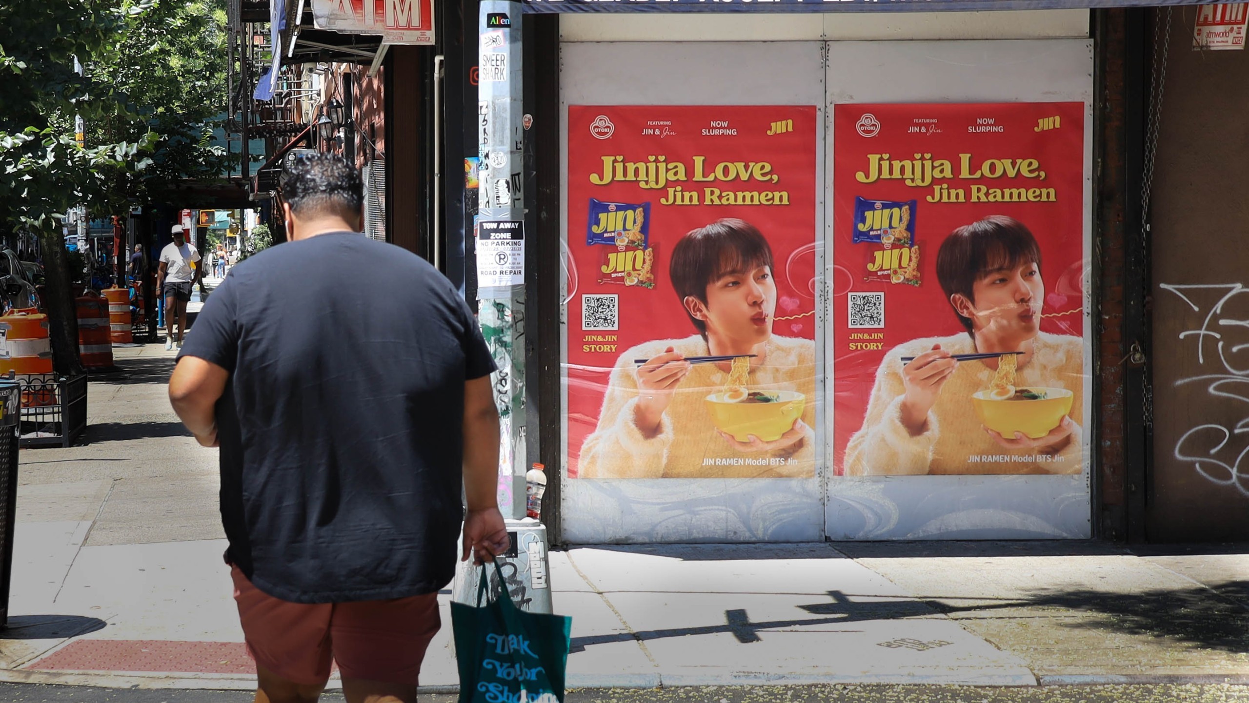 Jin Ramen Wild Posting® posters featuring BTS’s Jin displayed on a New York City storefront while a pedestrian walks by carrying a shopping bag.