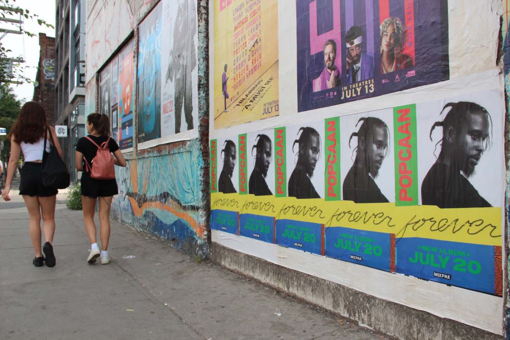 Wild Posting® street-level music poster installation on an urban wall, reaching pedestrians through repeated exposure in a high-traffic city corridor